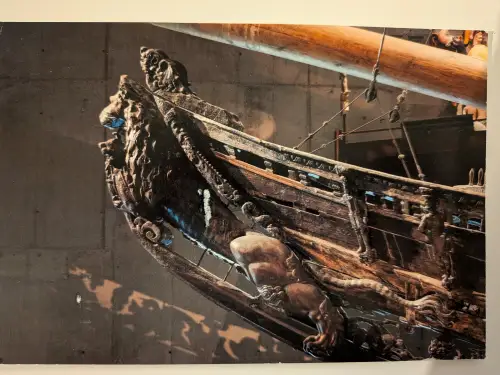 Ein Nahaufnahme des Bugs eines alten, dunklen Holzschiffs mit aufwendigen Schnitzereien und einer großen löwenähnlichen Figur vor dunklem Hintergrund.