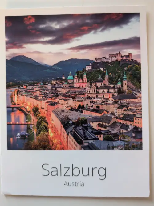 Eine Abendaufnahme von Salzburg zeigt die Festung Hohensalzburg über der beleuchteten Altstadt am Fluss Salzach unter einem farbigen Himmel.