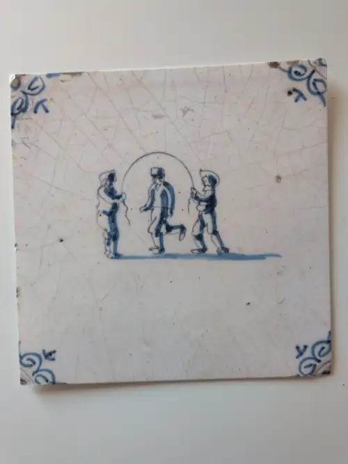 Eine alte, weiß-blaue Keramikfliese zeigt drei Figuren beim Seilspringen, umrahmt von Eckverzierungen und feinen Rissen.