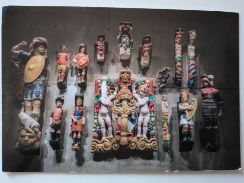 Eine Anordnung von farbig bemalten, geschnitzten Holzfiguren, darunter Krieger und Putten, ist auf einer dunklen Wand montiert.