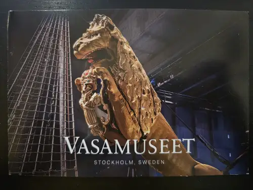 Eine große, goldene Löwenfigur mit offenem Maul und einem Wappen ist vor dunklem Hintergrund mit Schiffsrigg abgebildet, darunter der Text „VASAMUSEET STOCKHOLM, SWEDEN“.