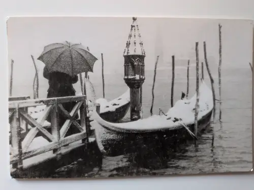 Ein Schwarz-Weiß-Bild zeigt schneebedeckte Gondeln, eine Person mit Regenschirm und einen verzierten Laternenpfahl in einem winterlichen Venedig.