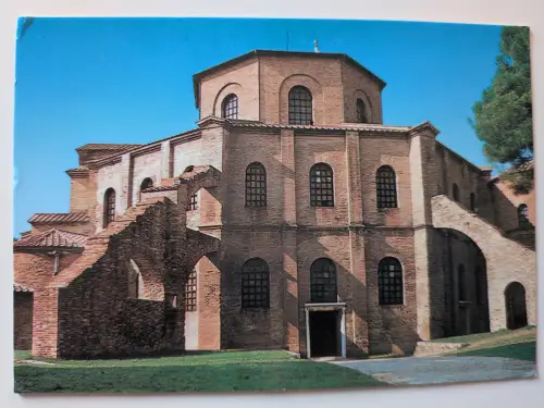 Ein großes, historisches Backsteingebäude mit achteckigem Kuppelbau und Rundbogenfenstern steht unter blauem Himmel auf grünem Gras.
