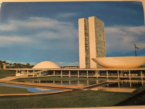 Das Bild zeigt den modernen Nationalkongress Brasiliens mit zwei hohen Türmen, einer Kuppel und einer Schale, umgeben von Wasserbecken und Grünflächen unter blauem Himmel.