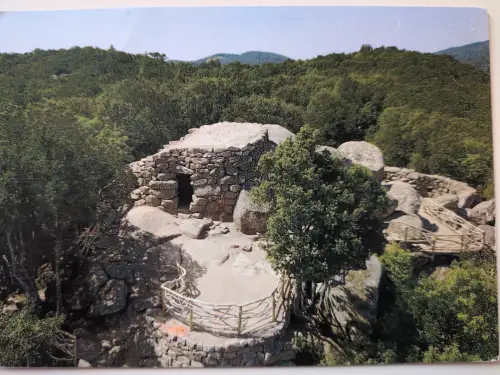 Eine alte Steinhütte mit dunklem Eingang steht auf felsigem Grund, umgeben von dichten Bäumen und Sträuchern unter einem klaren blauen Himmel.