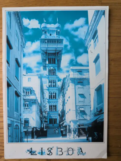 Eine blau getönte Postkarte zeigt den hohen, verzierten Elevador de Santa Justa in Lissabon, umgeben von Stadtgebäuden, mit dem Schriftzug „LISBOA“ am unteren Rand.