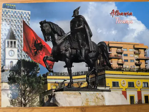 Das Bild zeigt eine Reiterstatue im Zentrum von Tirana, Albanien, umgeben von modernen Gebäuden, einem Uhrturm und der wehenden albanischen Flagge.