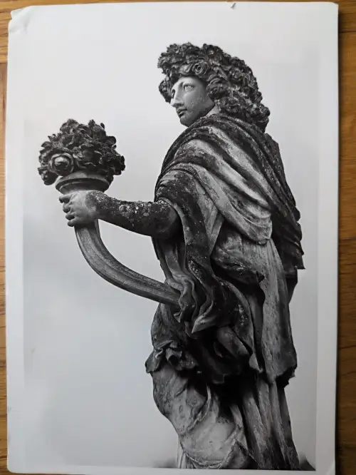 Eine verwitterte, schwarz-weiße Steinstatue zeigt eine Figur mit reicher Lockenpracht, die einen verzierten Kelch mit Blumen und kleinen Gesichtern in der Hand hält und nach links oben blickt.