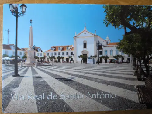 Ein sonniger Stadtplatz in Vila Real de Santo António mit einem markanten schwarz-weiß gestreiften Pflaster, einer weißen Kirche und einem Obelisken.