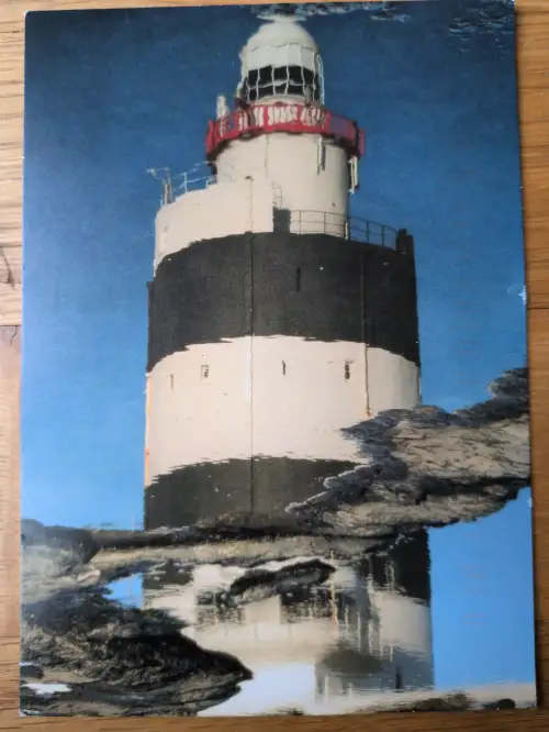 Ein schwarz-weiß gestreifter Leuchtturm mit rotem Band steht an einer felsigen Küste unter blauem Himmel, sein Spiegelbild im Wasser.