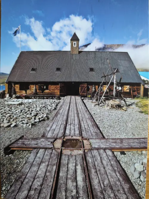 Ein großes, altes Holzgebäude mit einem Bootslip und einer wehenden Flagge steht vor einer Berglandschaft unter blauem Himmel.