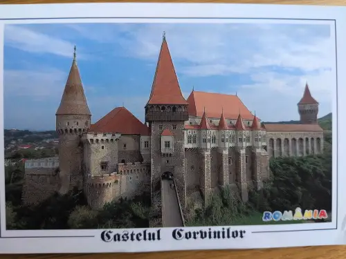 Ein imposantes, mittelalterliches Schloss mit zahlreichen Türmen und roten Dächern, verbunden durch eine lange Brücke, thront in grüner Landschaft.