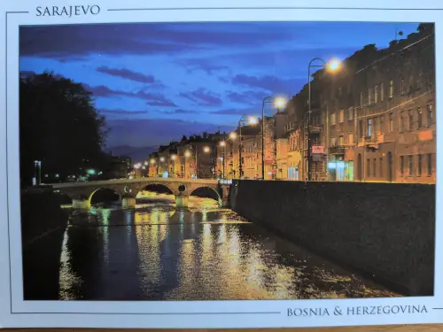 Eine nächtliche Stadtansicht von Sarajevo zeigt einen Fluss mit einer beleuchteten Bogenbrücke und hell erleuchteten Gebäuden am Ufer, deren Lichter sich im Wasser spiegeln.