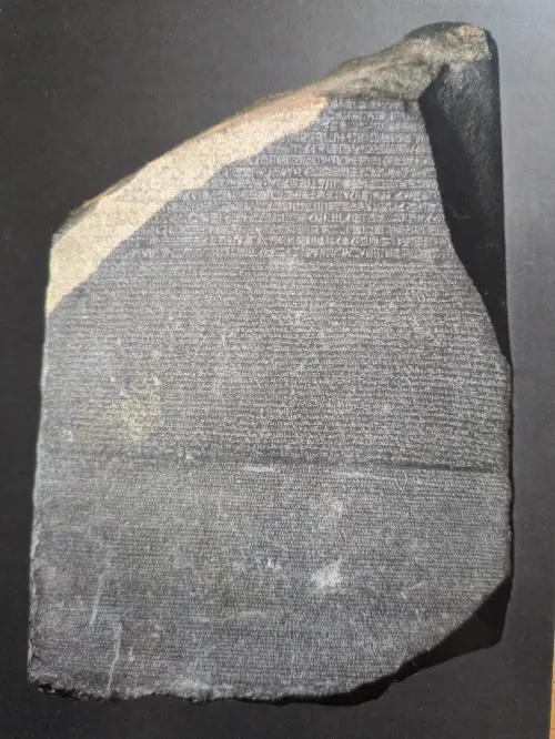 Eine dunkle, unregelmäßig geformte Steinplatte ist dicht mit verschiedenen antiken Schriften bedeckt. Es handelt sich um den Rosetta Stone.