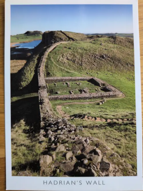 Der Hadrianswall, eine lange Steinmauer, zieht sich durch eine grüne Hügellandschaft mit einem See und antiken Ruinen unter blauem Himmel.