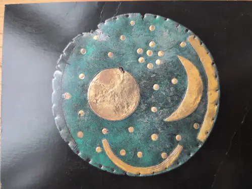 Eine runde, grünspanfarbene Bronzescheibe zeigt goldene Applikationen: einen großen Kreis in der Mitte, eine Mondsichel rechts, zahlreiche kleine Punkte als Sterne und einen Bogen am unteren Rand.