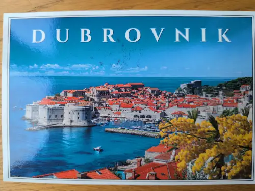Das Bild zeigt die historische Küstenstadt Dubrovnik mit roten Dächern, alten Mauern und einem Hafen am tiefblauen Meer, mit gelben Blüten im Vordergrund und dem Schriftzug "DUBROVNIK" oben.