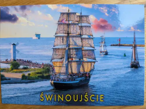 Ein großes Segelschiff fährt in Świnoujście auf dem Wasser, umgeben von weiteren Schiffen, einem Windmühlen-Leuchtturm und vielen Menschen am Ufer.