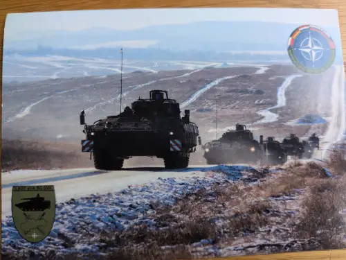 Eine Kolonne dunkler, gepanzerter Militärfahrzeuge fährt auf einer schneebedeckten Straße durch eine weite, hügelige Winterlandschaft, mit NATO- und Militärabzeichen im Bild.