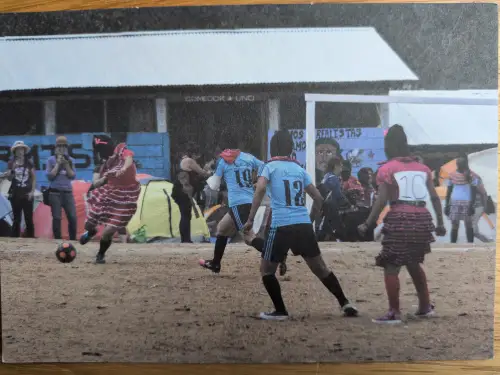 Mehrere Personen spielen Fußball auf einem staubigen Feld, einige in Sporttrikots, andere in farbigen Kleidern, mit Gebäuden und Zelten im Hintergrund.
