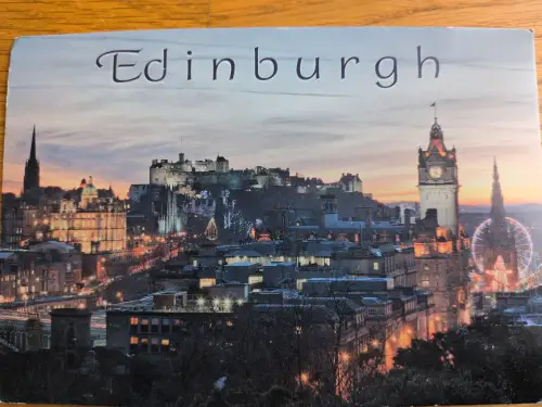 Ein nächtliches Stadtpanorama von Edinburgh mit dem beleuchteten Schloss, vielen Gebäuden und einem Riesenrad, oben steht "Edinburgh".