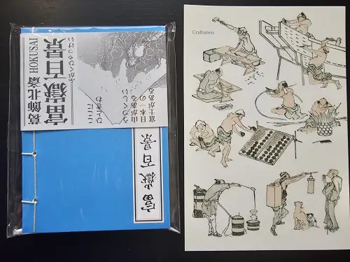 Ein blaues, in Folie verpacktes Büchlein mit japanischer Schrift und dem Namen Hokusai liegt neben einem Blatt mit farbigen Zeichnungen von Handwerkern bei der Arbeit.