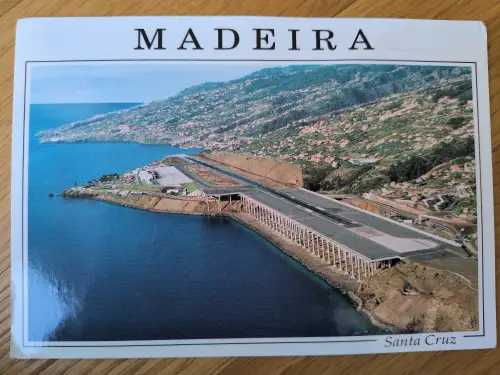 Das Bild zeigt eine Luftaufnahme des Flughafens von Madeira bei Santa Cruz, dessen Landebahn auf Pfeilern über dem tiefblauen Meer liegt, umgeben von grünen, dicht bebauten Berghängen.