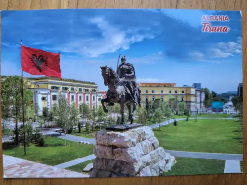 Ein Reiterstandbild steht in einem Park vor farbigen Gebäuden unter der wehenden albanischen Flagge in Tirana.