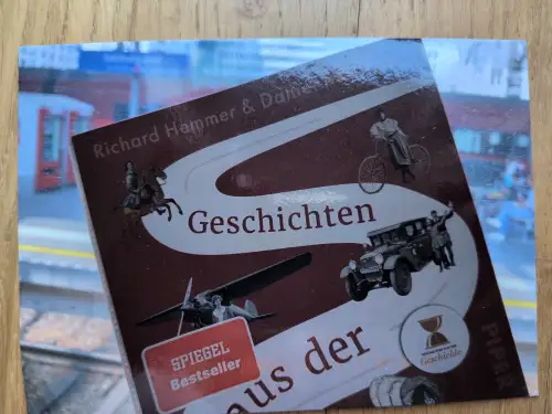 Im Vordergrund das Cover des Buches "Geschichten aus der Geschichte" von Richard Hemmer & Daniel Meßner zeigt historische Illustrationen von Reiter, Hochradfahrer, altem Auto und Flugzeug. Im Hintergrund ist verschwommen ein Bahnsteig zu sehen.