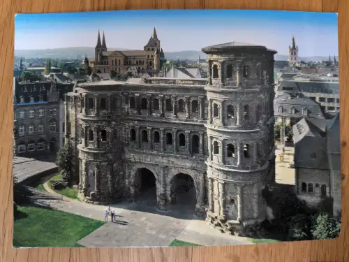 Die Porta Nigra, ein massives römisches Sandsteintor mit zwei Türmen und Bögen, steht im Zentrum des Bildes, dahinter eine Stadtlandschaft mit Kathedrale unter blauem Himmel.