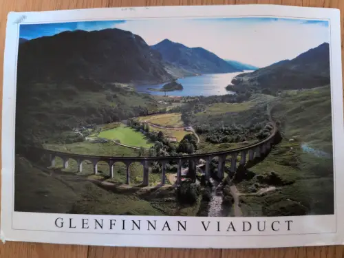 Ein Luftbild zeigt das geschwungene Glenfinnan Viadukt mit seinen Steinbögen in einer weiten grünen Berglandschaft mit einem See im Hintergrund.