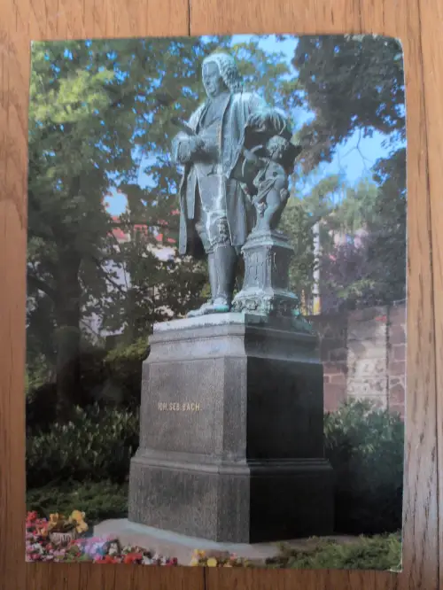Eine Bronzestatue von Johann Sebastian Bach steht auf einem hohen Steinsockel mit der Aufschrift "JOH. SEB. BACH.", umgeben von Bäumen und Blumen in einem Park.