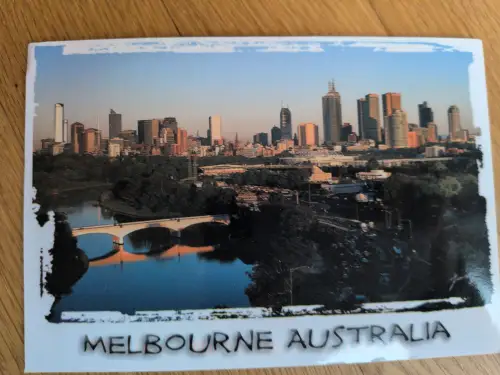Eine Postkarte zeigt die Skyline von Melbourne, Australien, mit einem Fluss, einer Bogenbrücke und grünen Ufern.