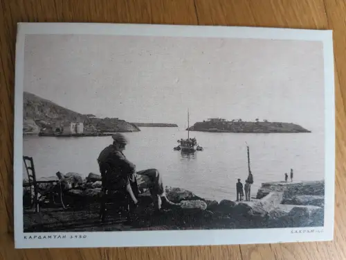 Schwarz-Weiß-Foto einer Bucht in Kardamyli (1930) mit einer sitzenden Person am Ufer, einem Boot auf dem Wasser und einer Insel im Hintergrund.