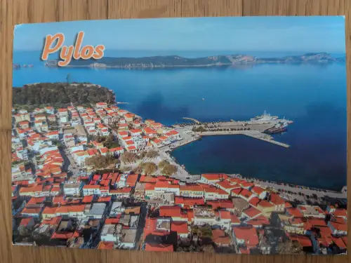 Eine Luftaufnahme zeigt die Küstenstadt Pylos mit dicht stehenden Häusern mit roten Dächern, einem Hafen mit Booten und dem weiten blauen Meer.