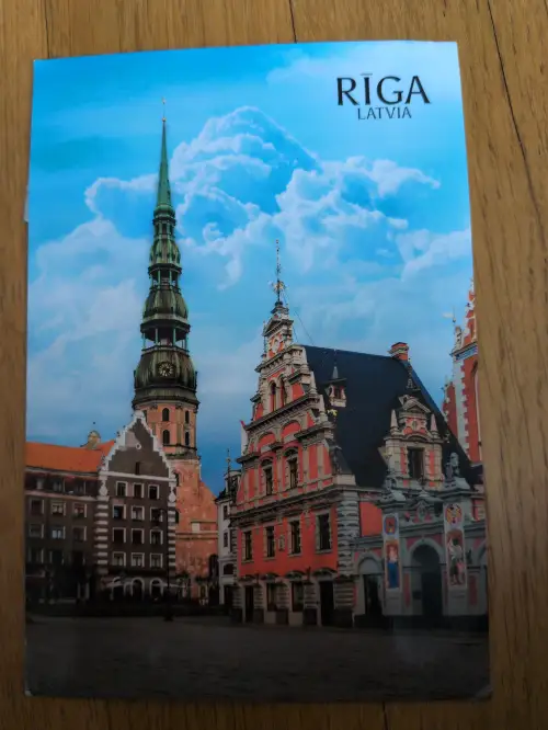 Das Bild zeigt eine historische Stadtansicht von Riga mit einem hohen grünen Kirchturm und farbenfrohen Giebelhäusern unter einem blauen Himmel mit Wolken.