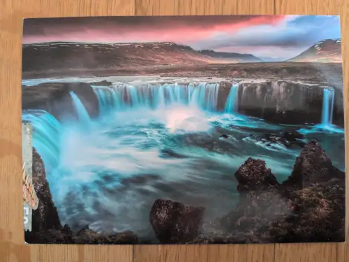 Ein breiter Wasserfall mit leuchtend blauem Wasser stürzt in eine felsige Schlucht, umgeben von Bergen unter einem rosa-blauen Himmel.