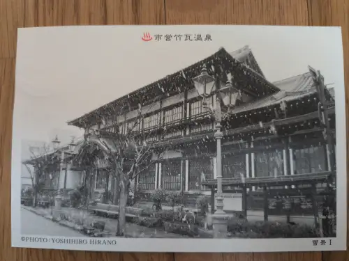 Ein Schwarz-Weiß-Foto zeigt ein großes, traditionelles japanisches Gebäude mit geschwungenen Dächern im Schneefall, umgeben von Bäumen und Laternen.