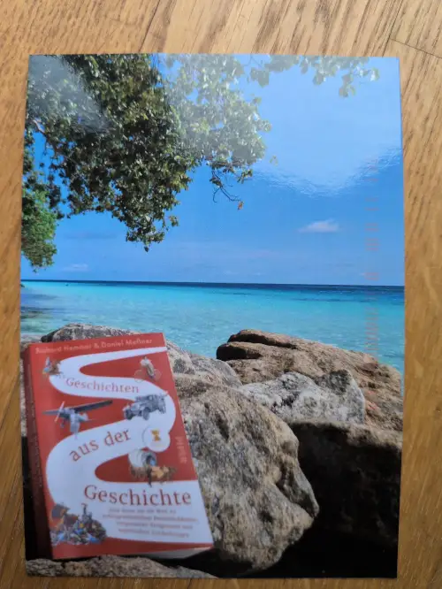 Ein rotes Buch mit dem Titel „Geschichten aus der Geschichte“ liegt auf Felsen an einem tropischen Strand mit blauem Meer und Himmel.