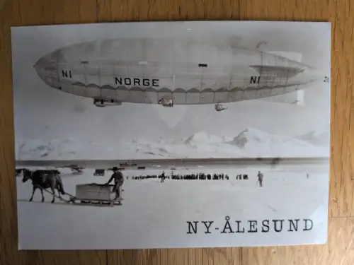 Ein Schwarz-Weiß-Foto zeigt das Luftschiff NORGE über dem verschneiten Ny-Ålesund, mit Menschen und einem Pferdeschlitten auf dem Eis.