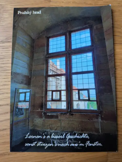Ein Innenraum mit Steinmauern und einem großen, mehrteiligen Holzfenster, das den Blick auf andere Gebäude und blauen Himmel freigibt.