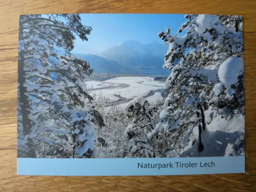 Eine winterliche Landschaft mit schneebedeckten Bäumen, die ein verschneites Flusstal und Berge unter blauem Himmel einrahmen, mit dem Schriftzug „Naturpark Tiroler Lech“ am unteren Bildrand.