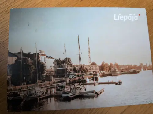 Das Bild zeigt eine Hafenansicht von Liepāja mit zahlreichen Segelbooten, Gebäuden am Ufer und einem hohen Sendeturm unter hellem Himmel.