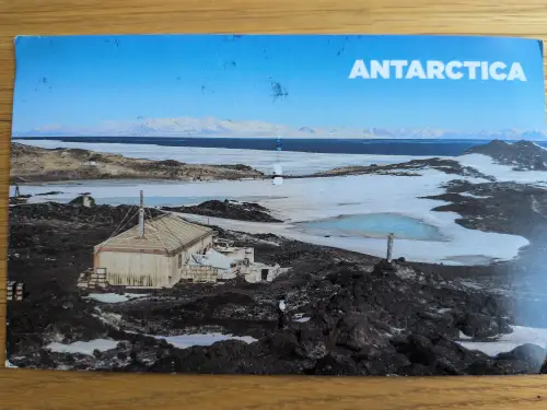 Ein Holzgebäude steht in einer eisigen Antarktislandschaft mit schneebedeckten Bergen im Hintergrund.