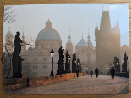 Eine historische Steinbrücke mit Statuen und beleuchteten Laternen, auf der Menschen im warmen, diffusen Licht vor einer Stadtkulisse mit Kuppeln und Türmen entlanggehen.