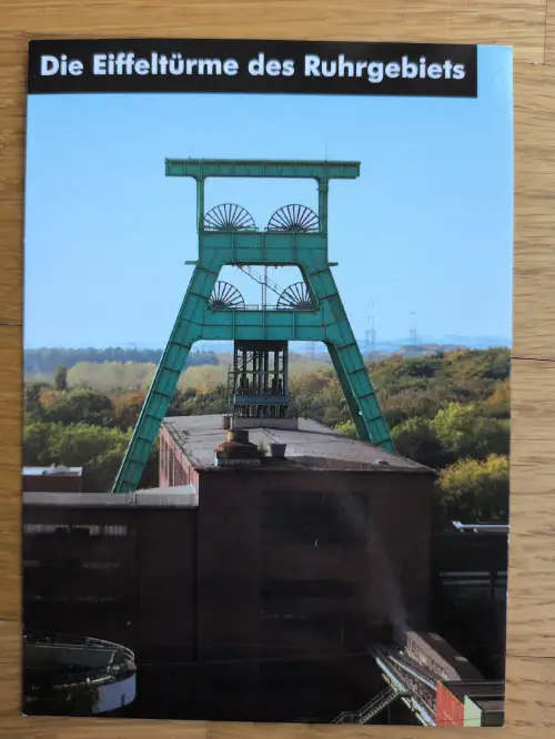 Das Bild zeigt einen grünen Förderturm mit großen Seilscheiben über einem Zechengebäude vor blauem Himmel, betitelt mit "Die Eiffeltürme des Ruhrgebiets".