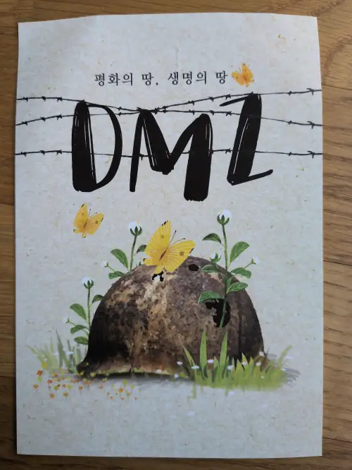 Ein Plakat zeigt die Buchstaben DMZ hinter Stacheldraht, darunter einen alten Militärhelm, aus dem Blumen sprießen und um den gelbe Schmetterlinge fliegen, mit koreanischem Text „Land des Friedens, Land des Lebens“ oben.