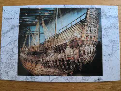 Das Bild zeigt das große, reich verzierte Holzschiff Vasa in einem Museum, umgeben von einer alten Seekarte.