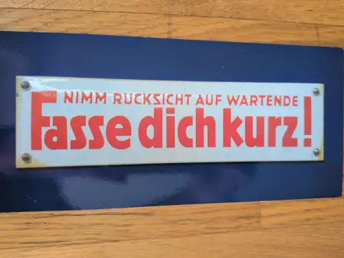 Ein weißes Schild mit roter Schrift auf blauem Untergrund zeigt die Aufforderung: "Nimm Rücksicht auf Wartende, Fasse dich kurz!".