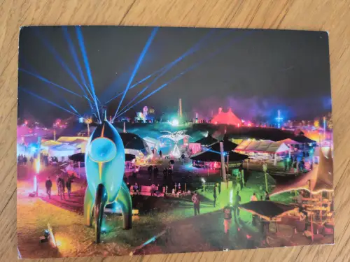 Ein farbenfrohes Nachtfestival mit einer großen, hellblauen Raketenskulptur im Vordergrund, umgeben von Zelten, Menschen und blauen Laserstrahlen am dunklen Himmel.